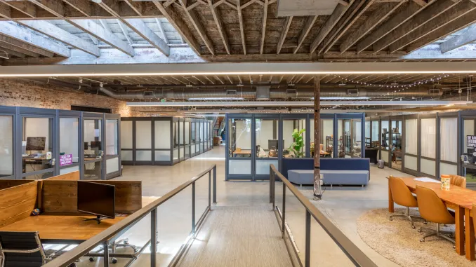 An airy workspace with glass-enclosed offices, exposed timber beams, brick walls and shared seating